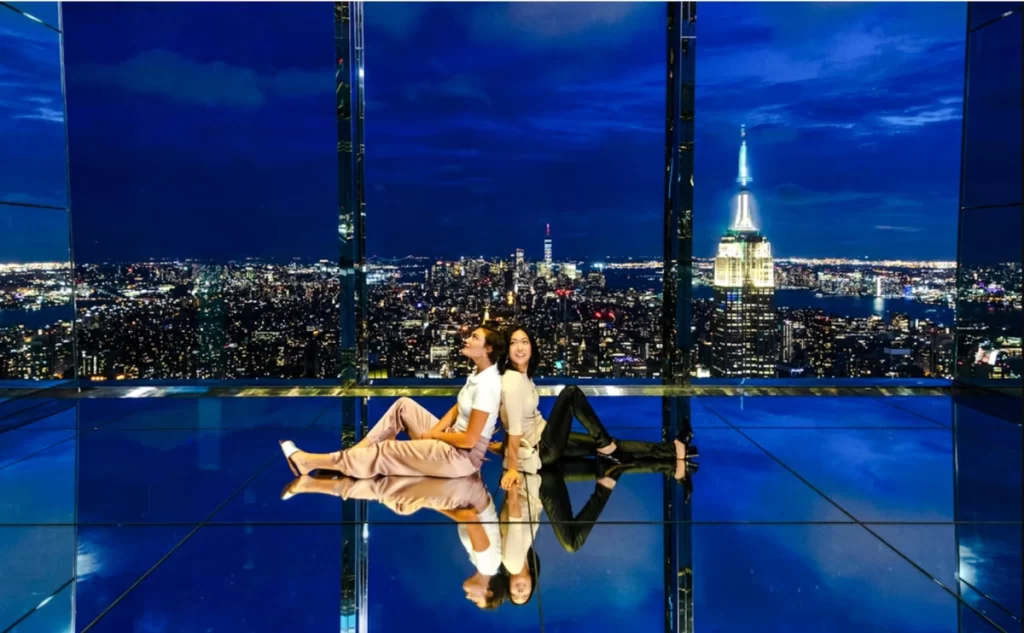 Visitantes sentados no chão espelhado com vista noturna do Empire State Building.