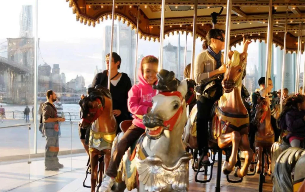 Crianças e adultos no Jane's Carousel, com a Ponte do Brooklyn e o skyline de Manhattan ao fundo.
