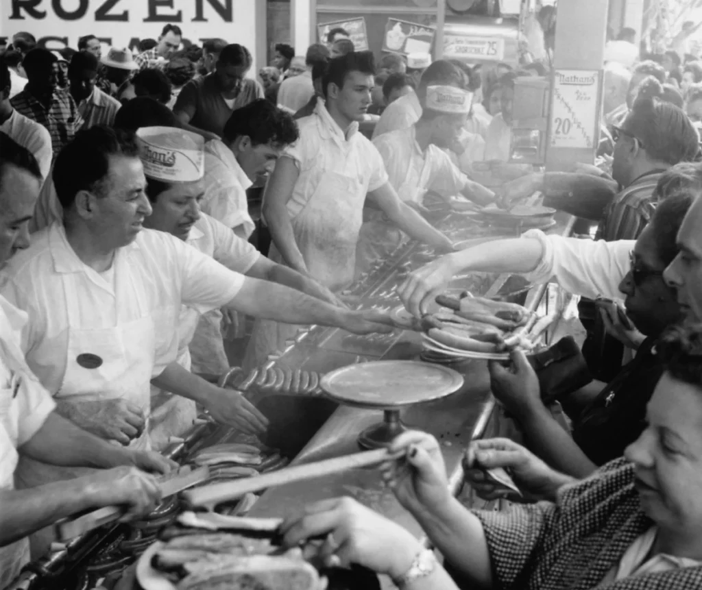 Fotografia histórica em preto e branco mostra funcionários servindo hot dogs a uma multidão no Nathan's Famous.