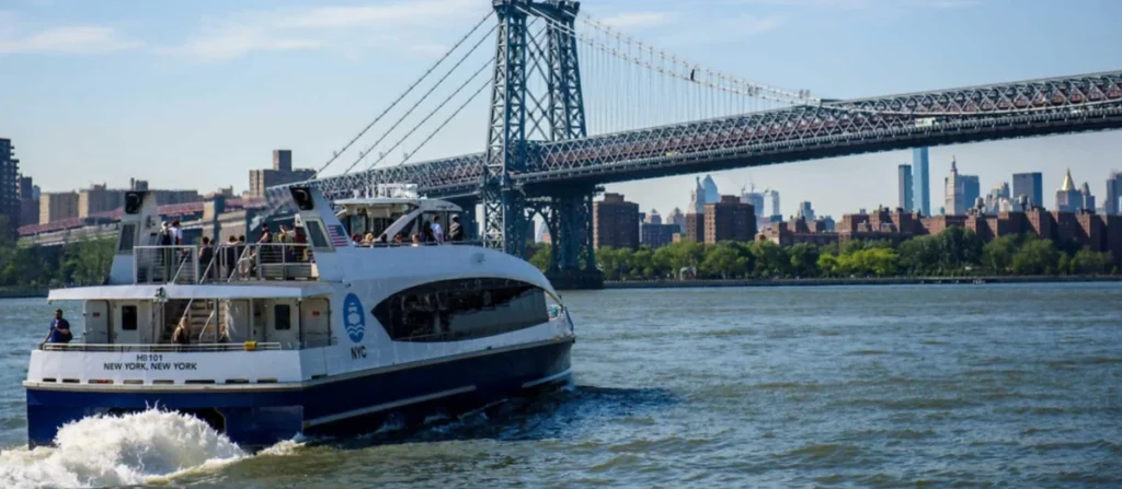 Ferry da NYC Ferry navegando pelo East River, com a Williamsburg Bridge e Manhattan ao fundo.