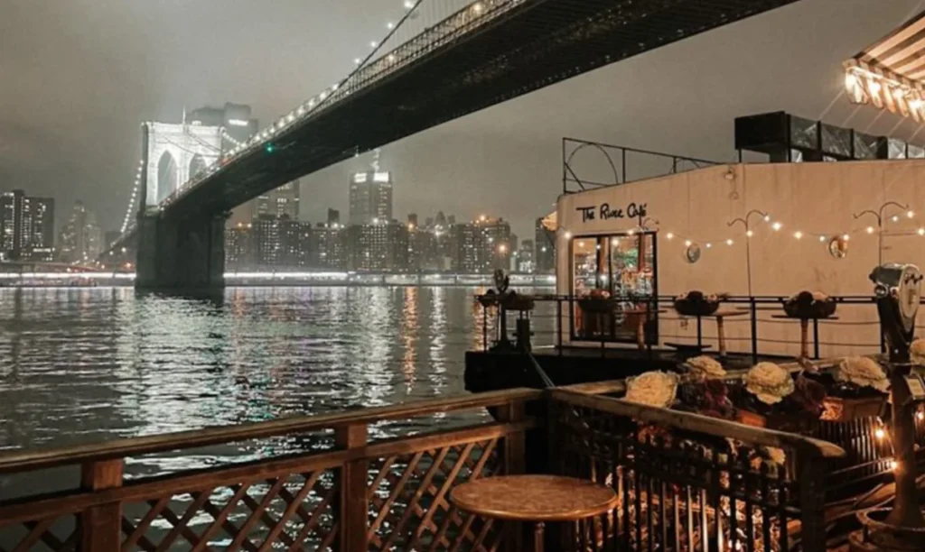 Vista noturna do The River Café à beira do East River, sob a Brooklyn Bridge e com Manhattan iluminada ao fundo.